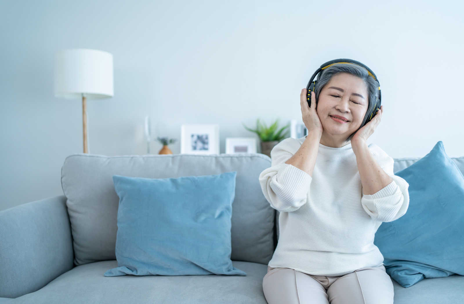 A senior woman smiling while relaxing on a couch and listening to music through wireless headphones.