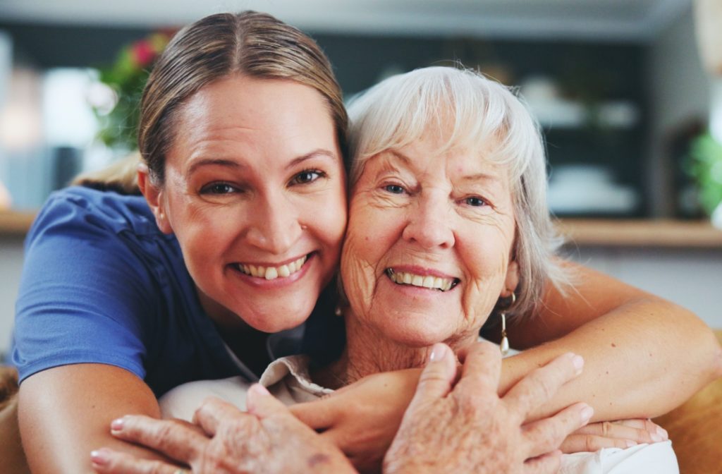 A caregiver hugging an older adult from behind while both smile in assisted living.