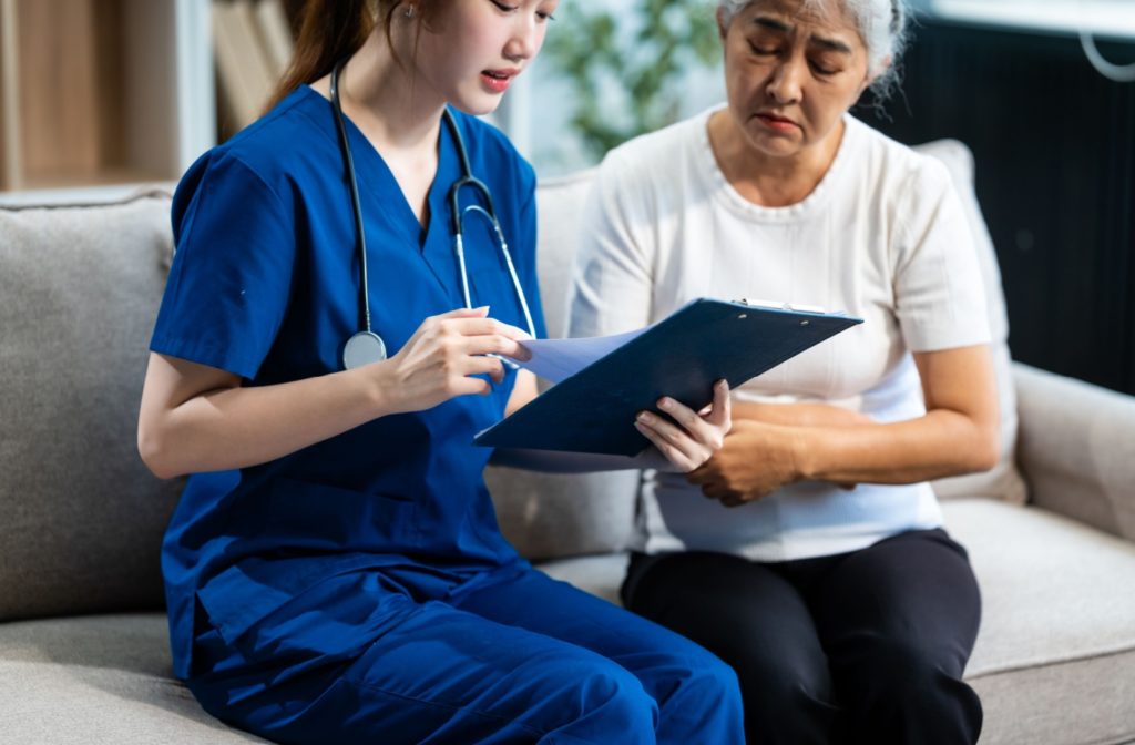 A caregiver in scrubs discussing a document with an older woman on a couch.