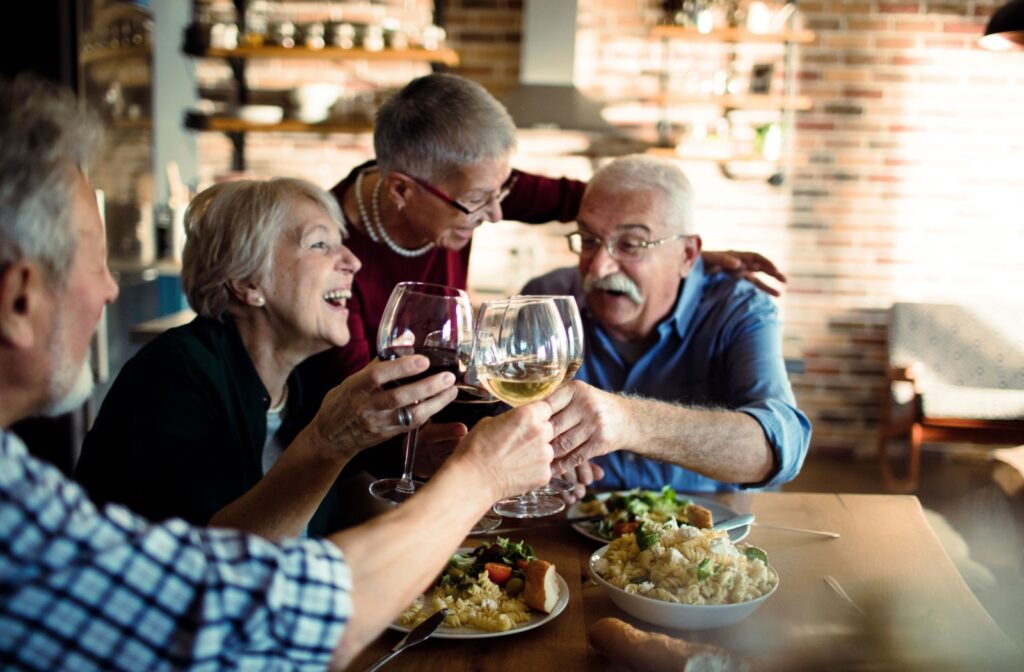 A group of seniors laugh while clinking their drinks together to celebrate delicious food in their senior living community.