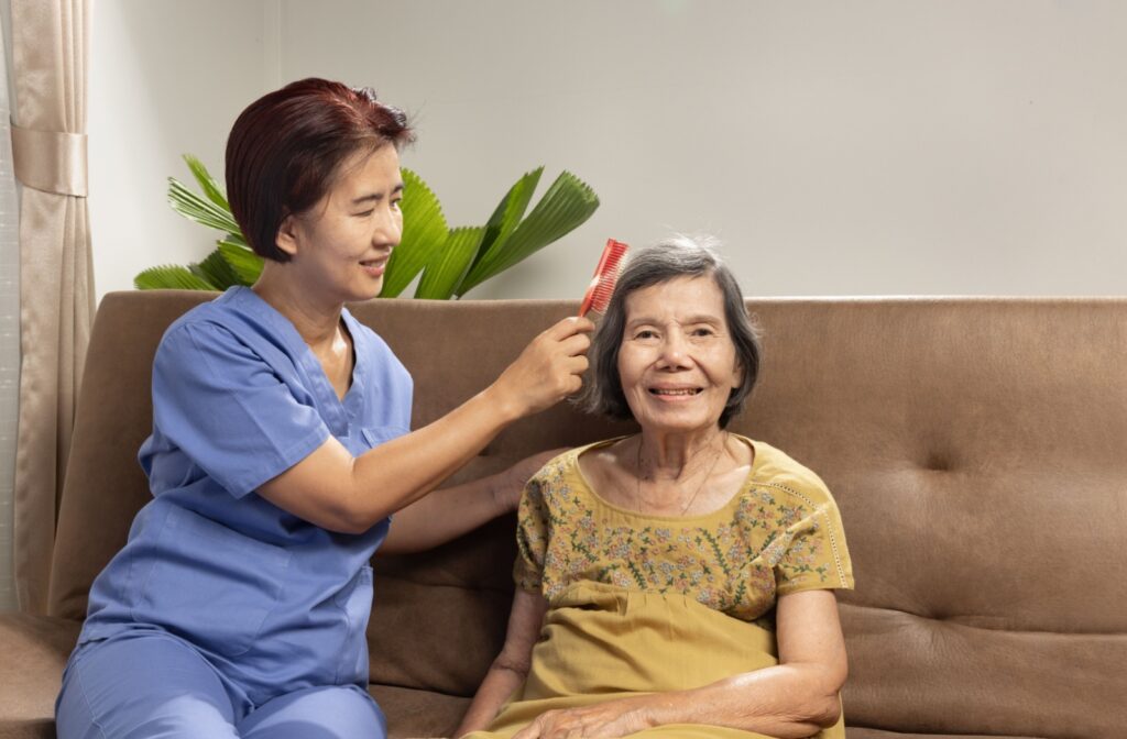 A caregiver carefully brushes a senior’s hair while seated on the couch to help them with personal grooming.