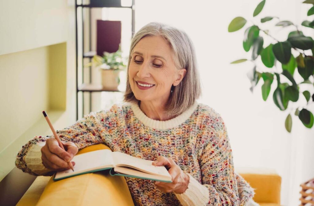 Older adult woman smiling in a rainbow coloured stitched sweater while writing in a journal in a cozy sunlit living room