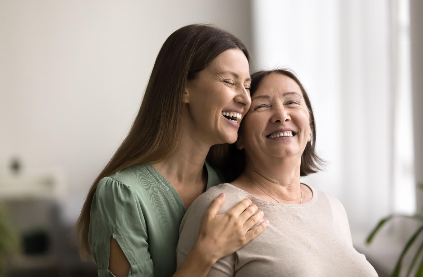 A person embraces their older parent from behind as they share a laugh.