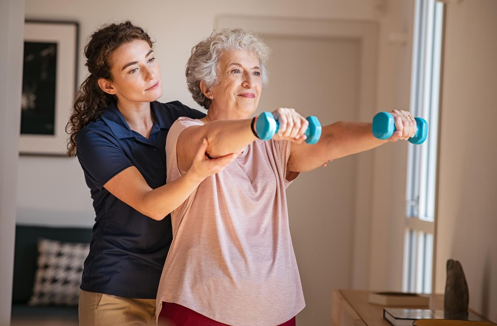 A caregiver carefully helping an older adult lift weights in senior living to help manage her chronic pain.