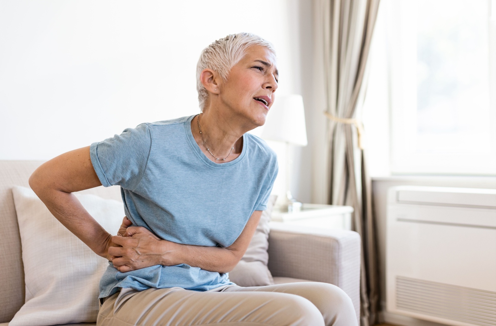 A senior wearing a light blue t-shirt sitting on a sofa in a living room holding their hip expressing pain.
