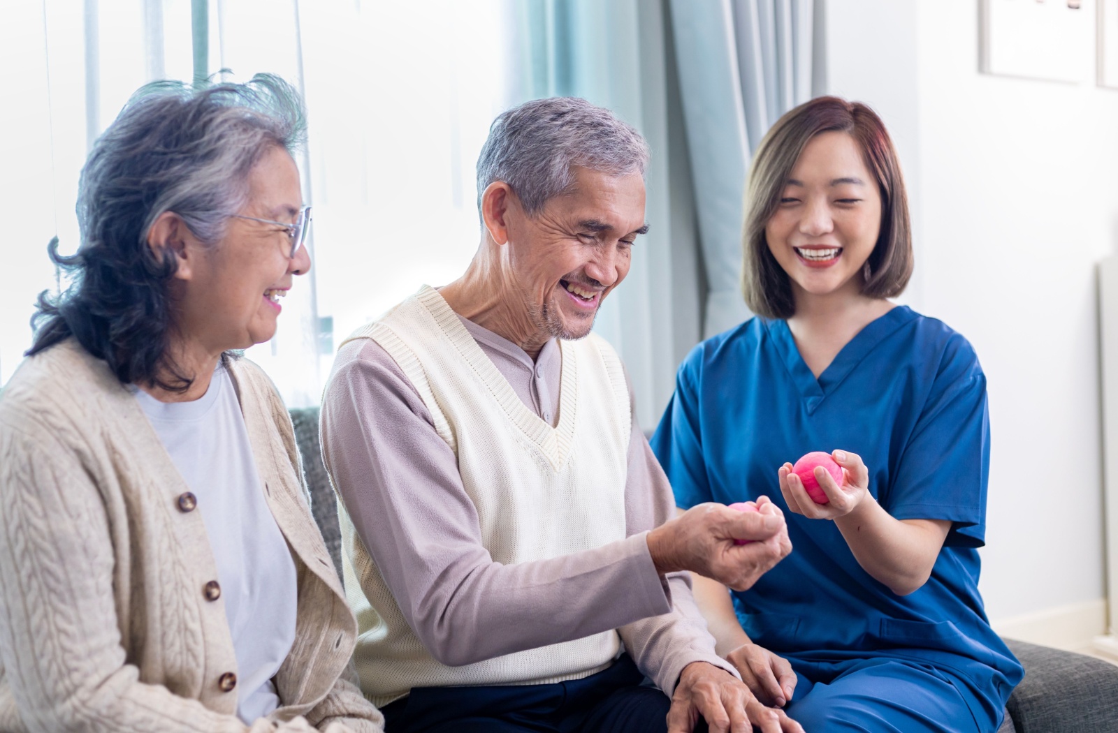 A healthcare worker in blue scrubs assisting two seniors with hand exercises using small therapy balls in a bright room.