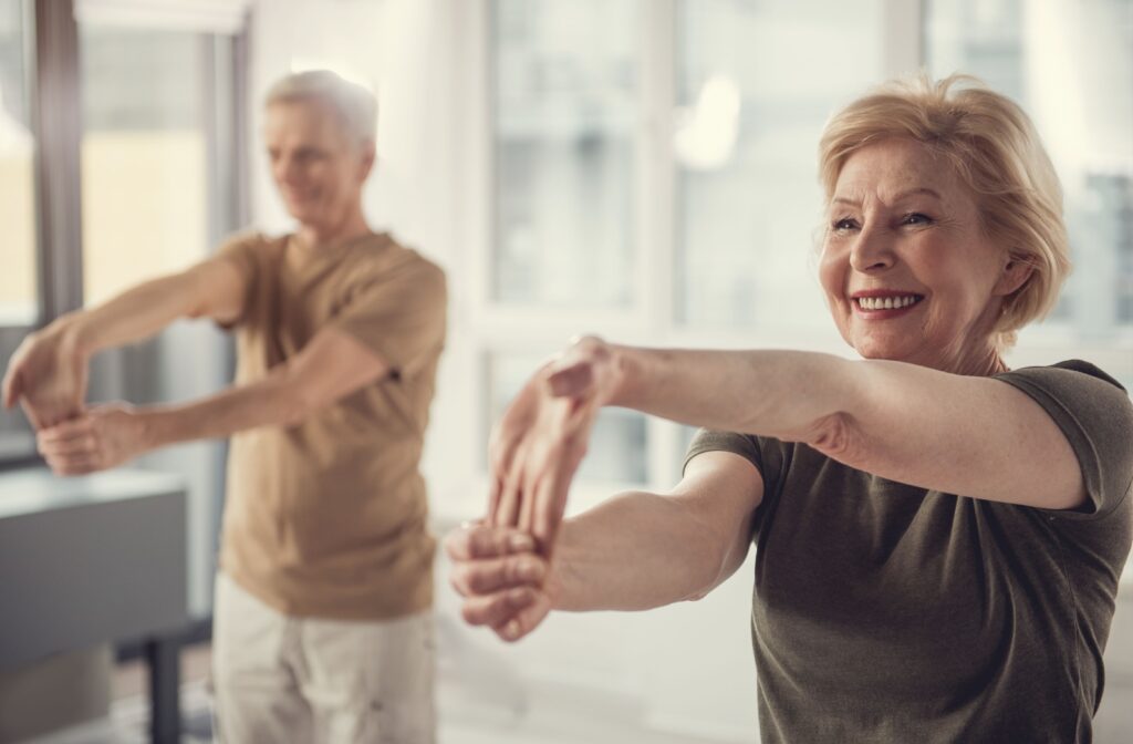 A smiling older adult in a green shirt stretching their wrists forward while another senior in a brown shirt follows along.