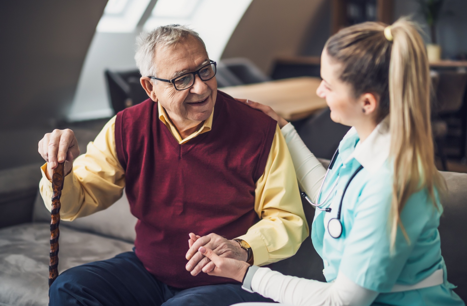 A caregiver smiling and holding hands with an older man sitting with a walking cane.