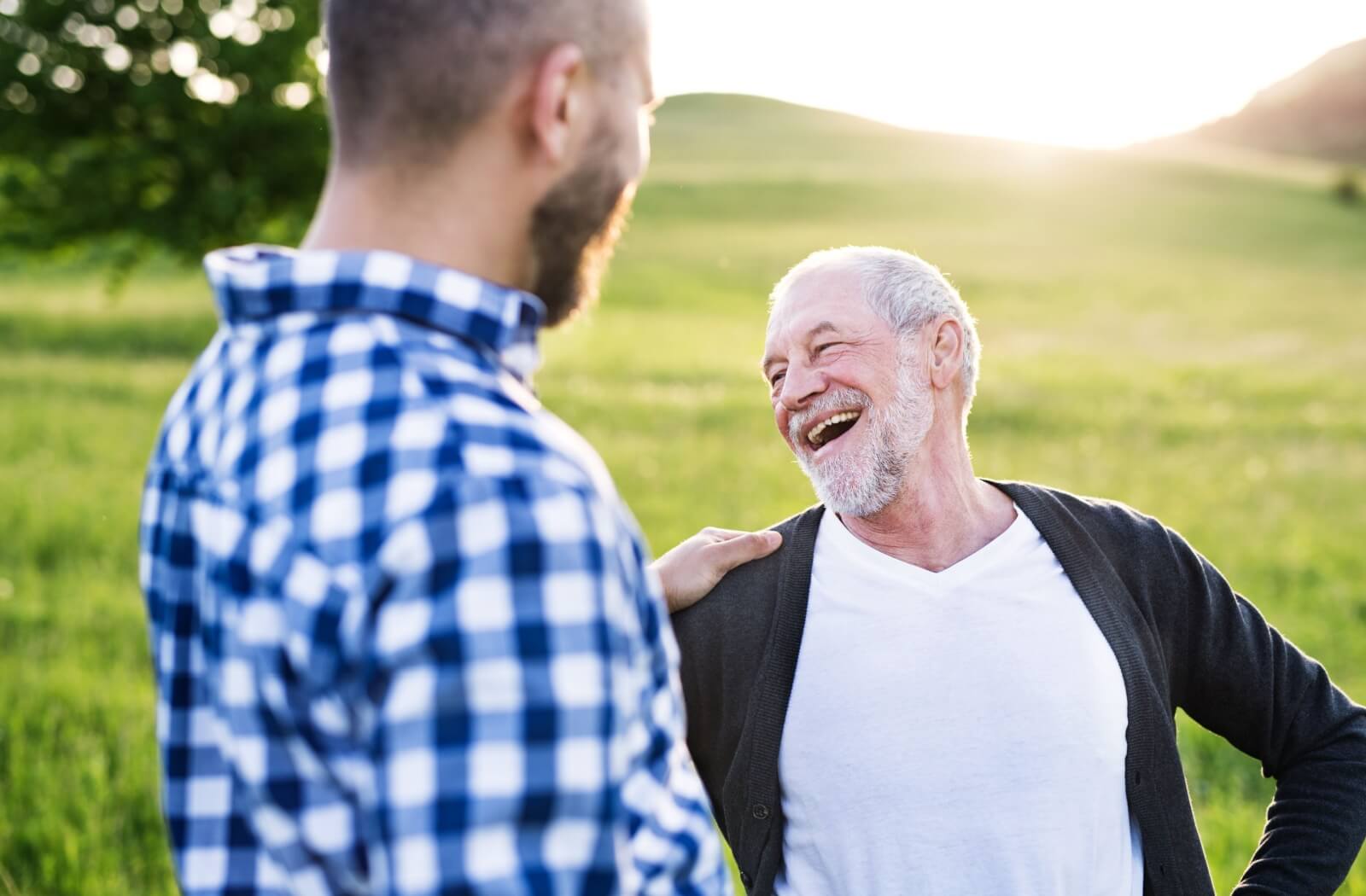 A senior and their adult child outdoors in a green field on a bright sunny day laughing and sharing in a moment of joy