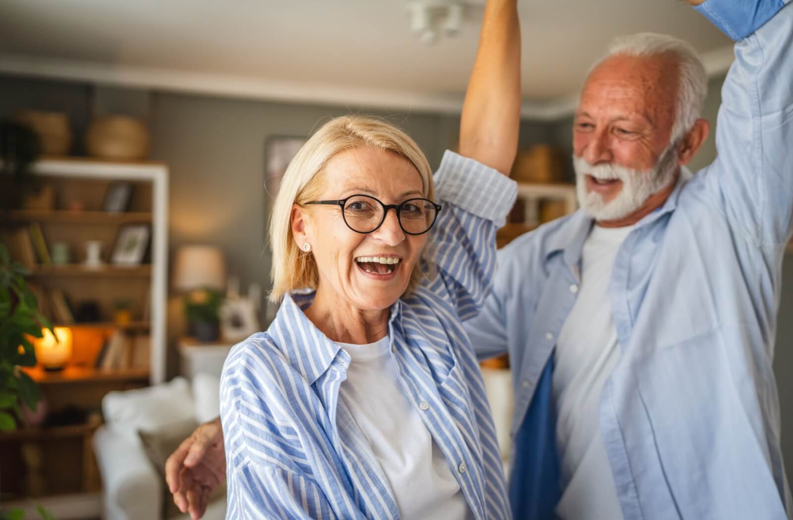 A senior couple wearing light blue button-up shirts dance together in a cozy living room environment, smiling and laughing.