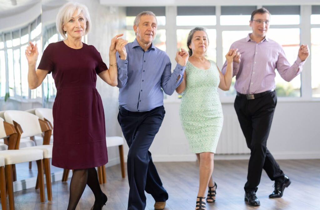 A group of 4 older adults, dressed in semi-formal clothing, hold hands and smile while performing a line dance in a bright room.