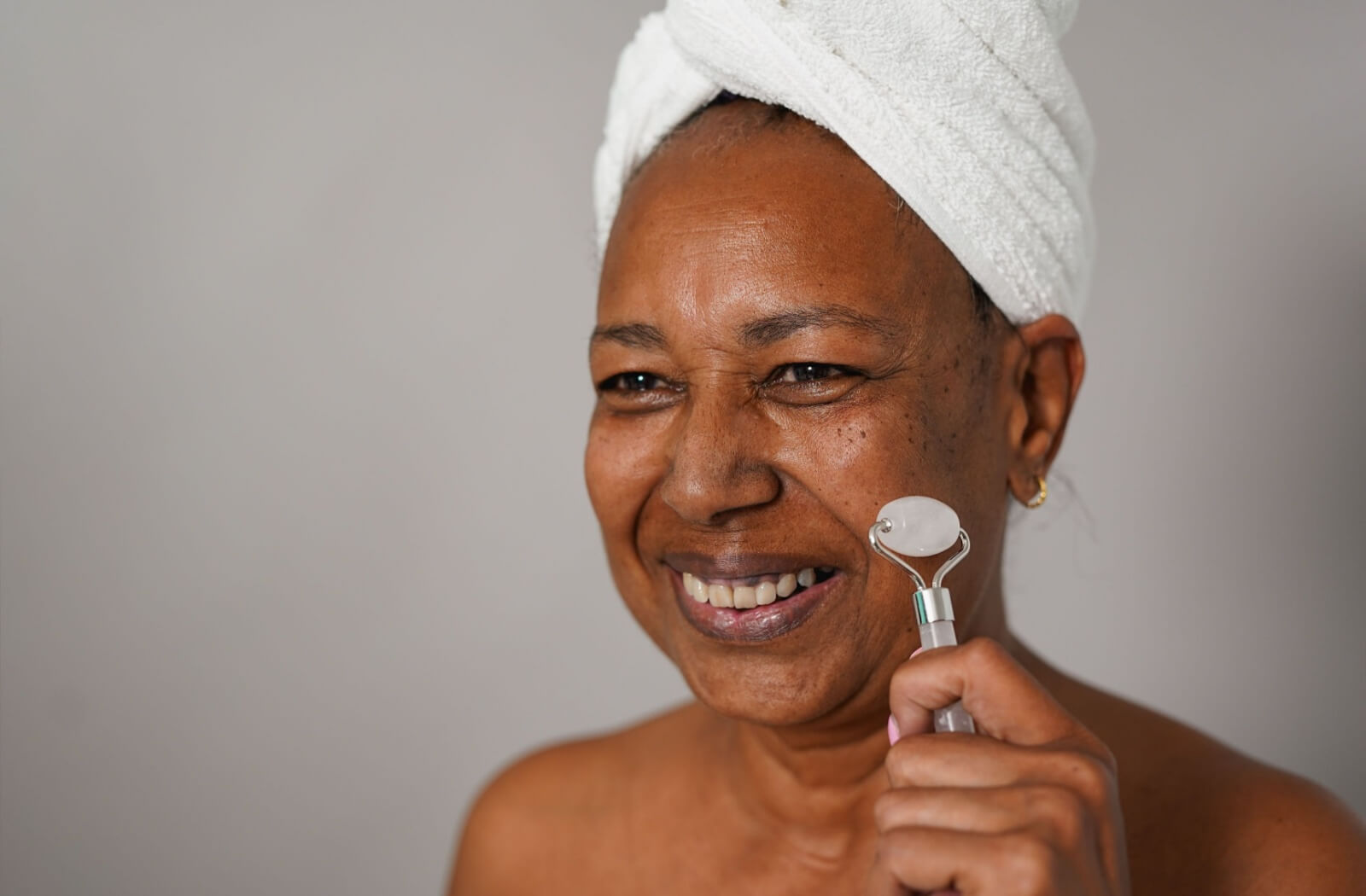 Smiling senior using a jade roller on face with towel wrapped around head promoting skin care and aging gracefully routines