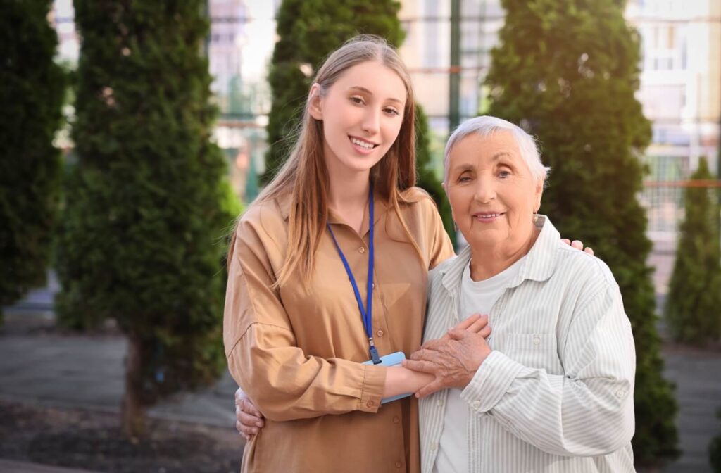 Young caregiver standing outside with their arm around an older adult as they smile together in a garden