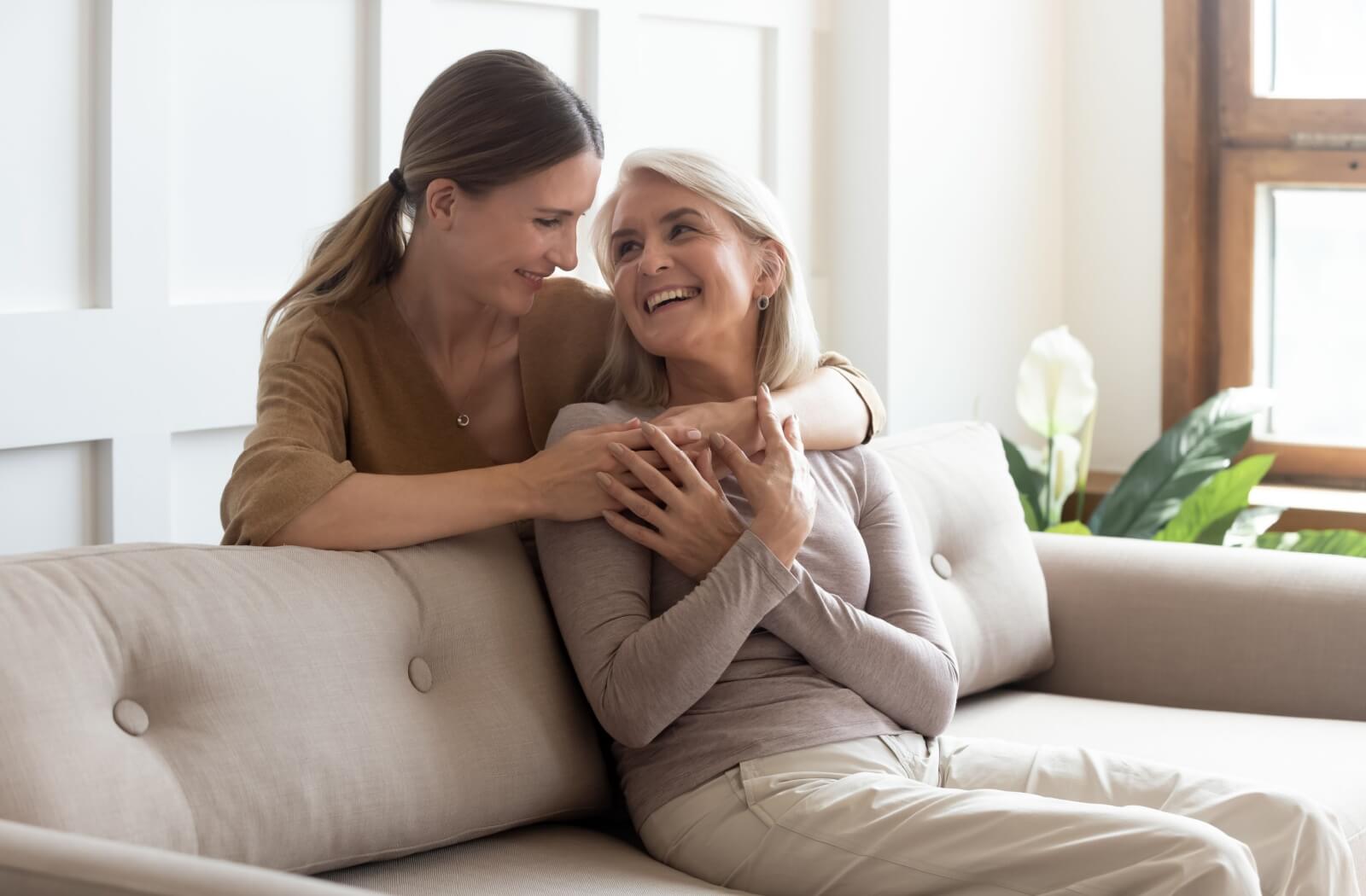An adult hugs their older parent over the shoulders to show love and affection during a visit to their senior living home