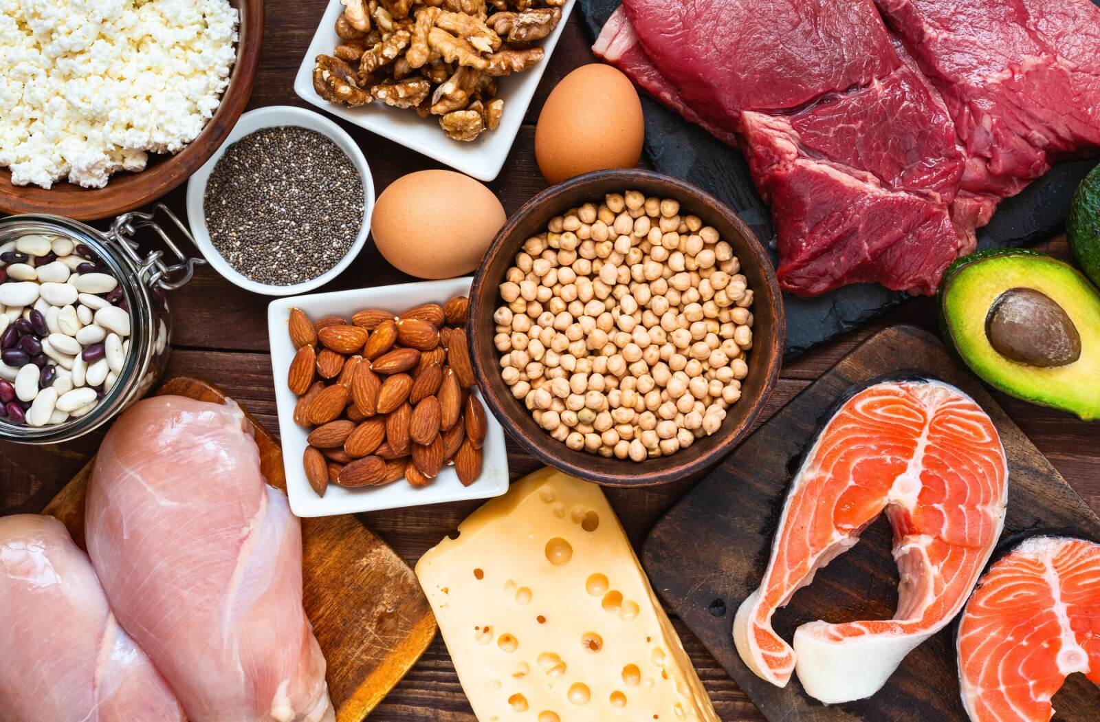 A wooden table full of various colorful types of nutritious foods, including fish, avocado, cheese, chickpeas, eggs, and nuts