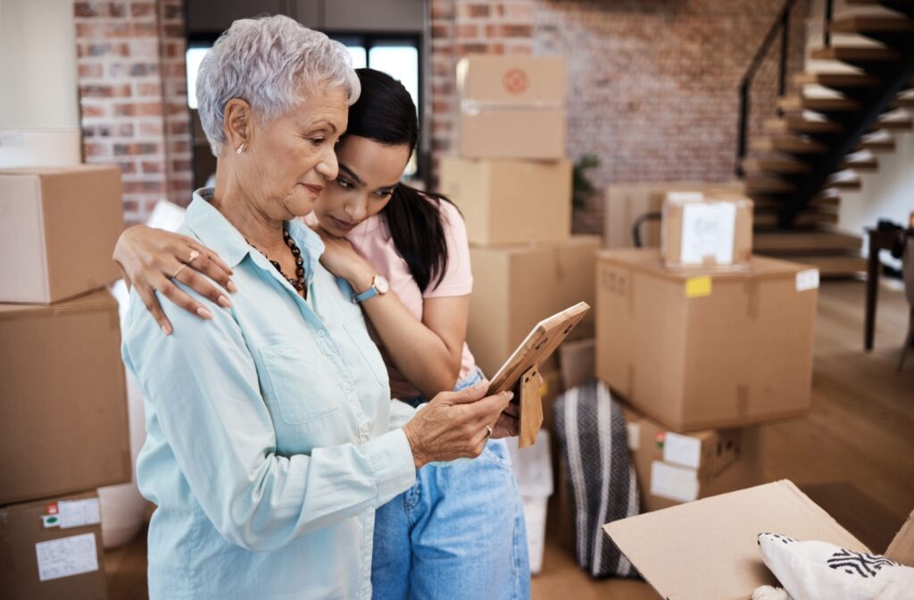 A mature adult reminisces over an old photograph with a family member while packing for their assisted living move.