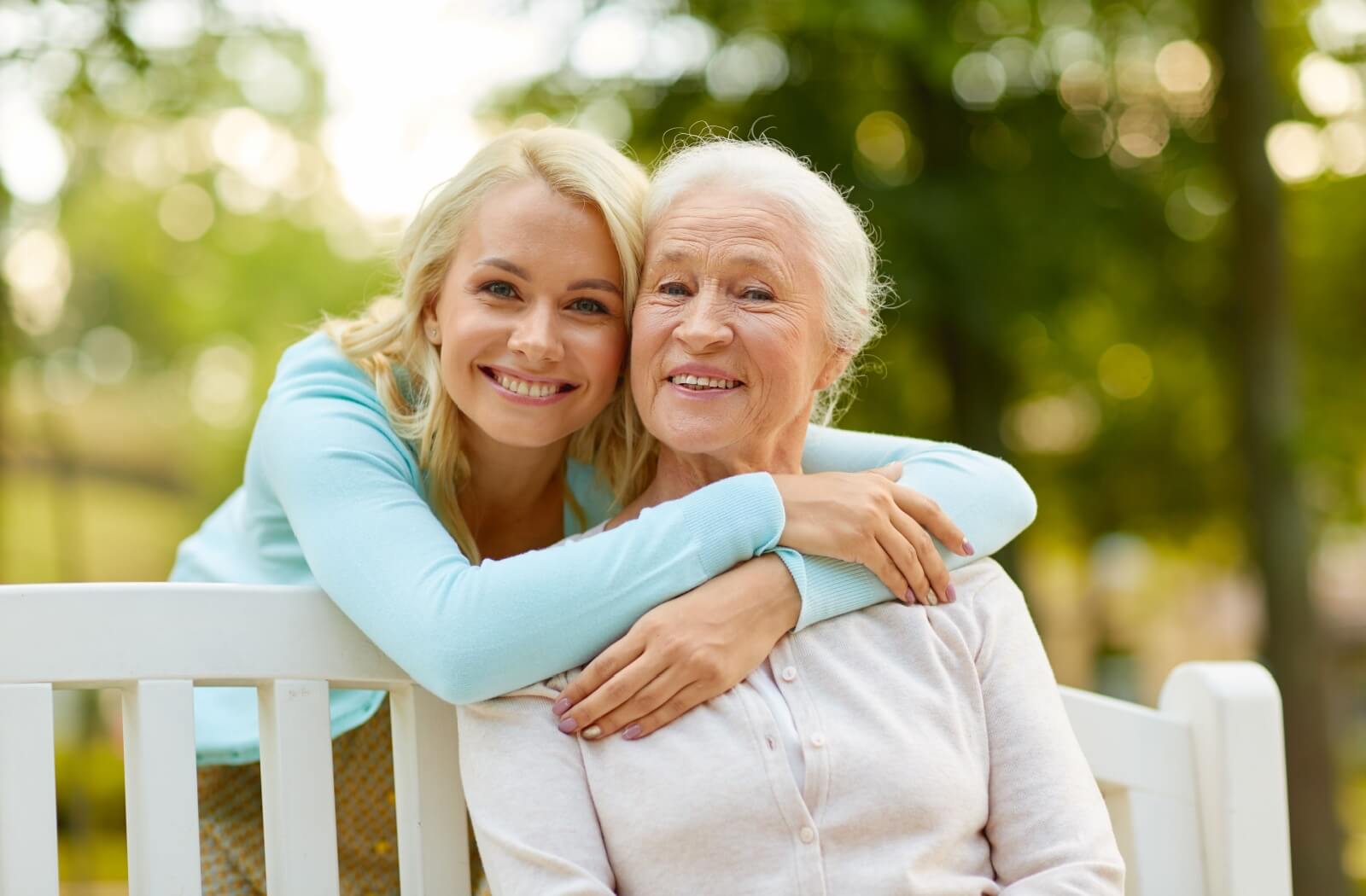 Younger adult smiling with arms around older adult on a park bench both smiling with greenery in the background