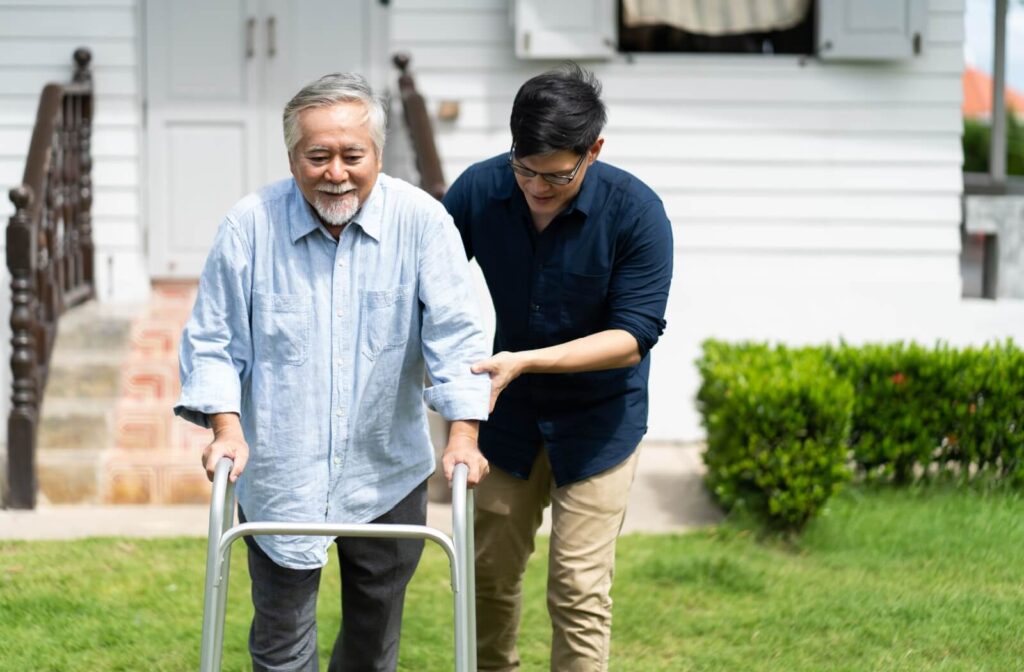 An older adult uses a walker assisted by a younger adult, enjoying fresh air outside a white house.
