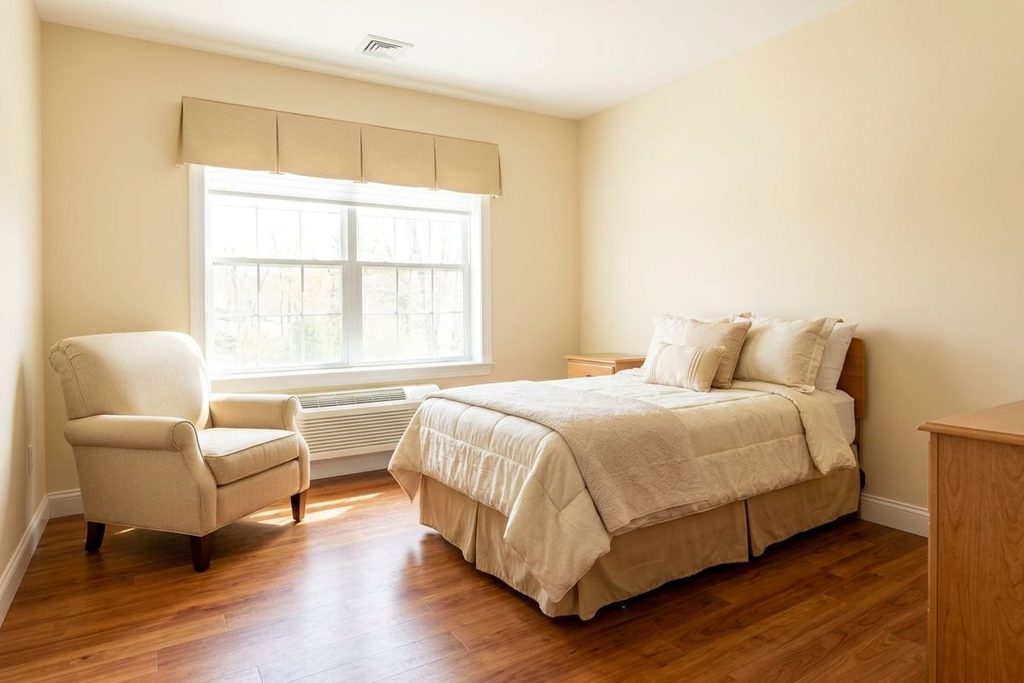 A bedroom is occupied by an upholstered armchair sitting by a large window and a bed in cream-colored bedding.